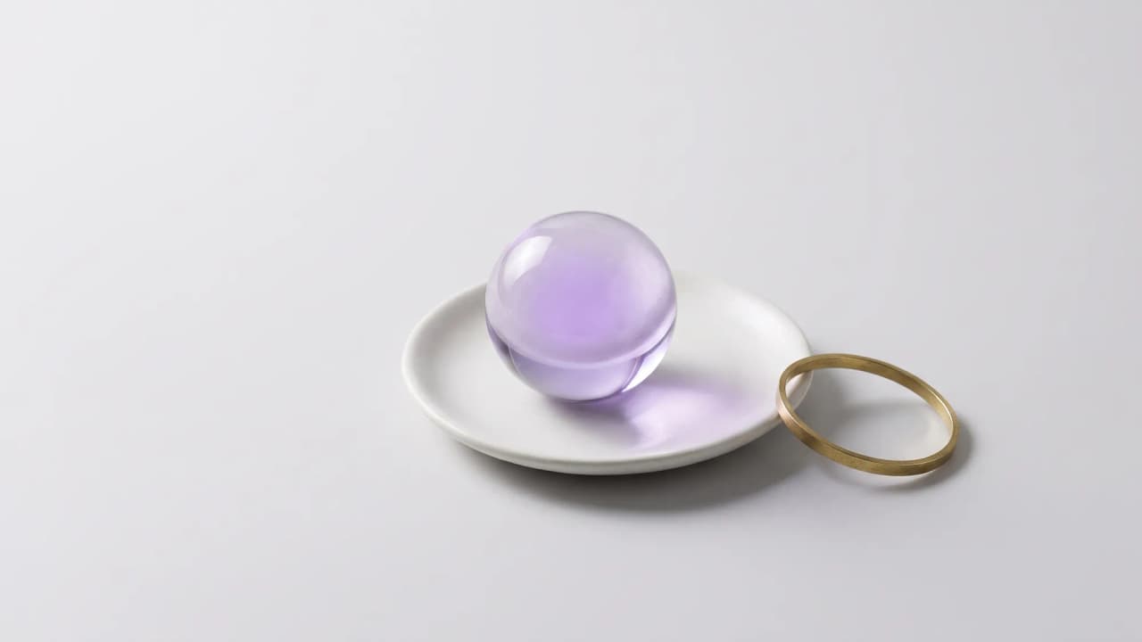 A clear glass sphere with a soft violet glow, on a small white dish beside a thin brass ring